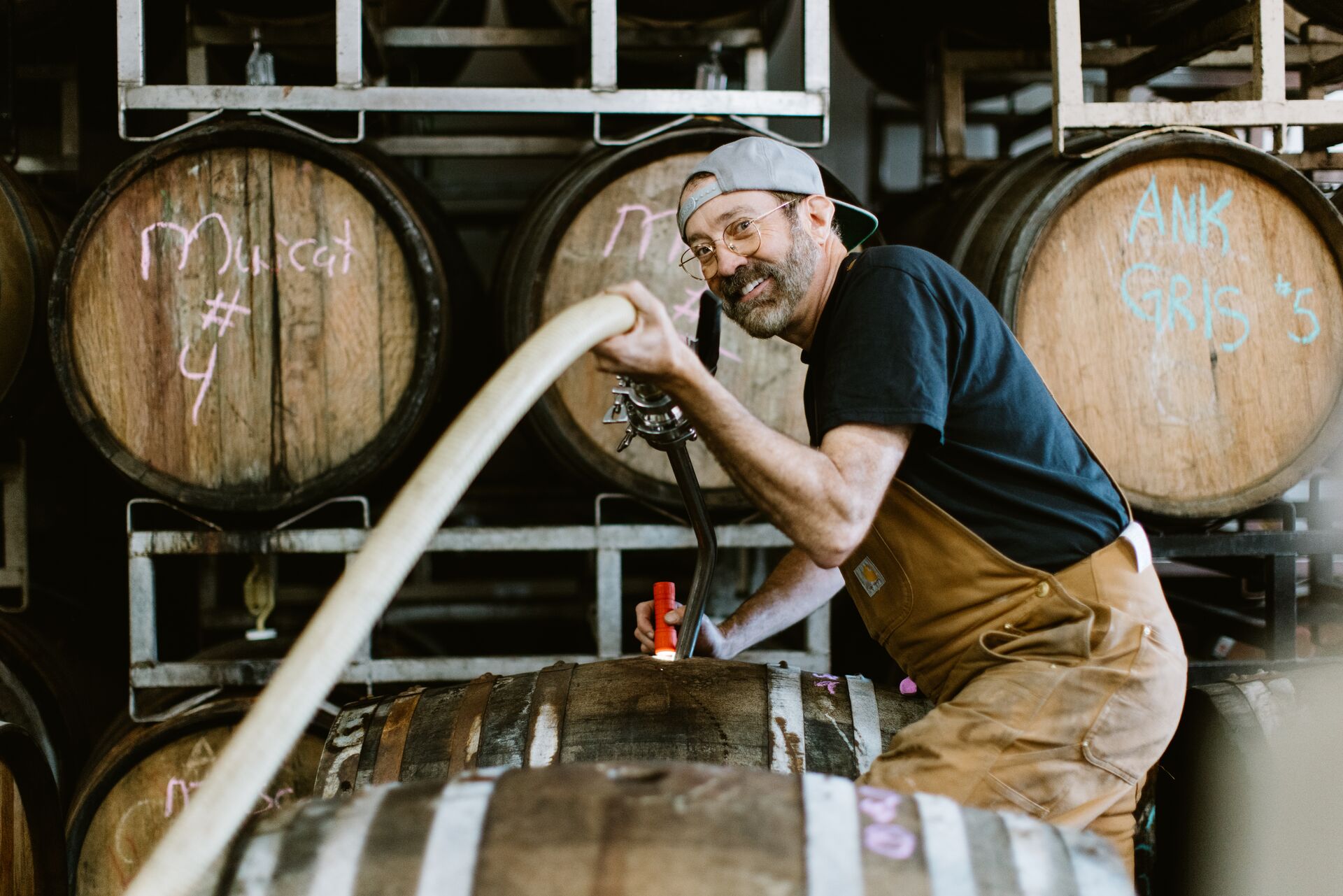 A smiling winemaker filling an oak barrel with a hose in the barrel room
