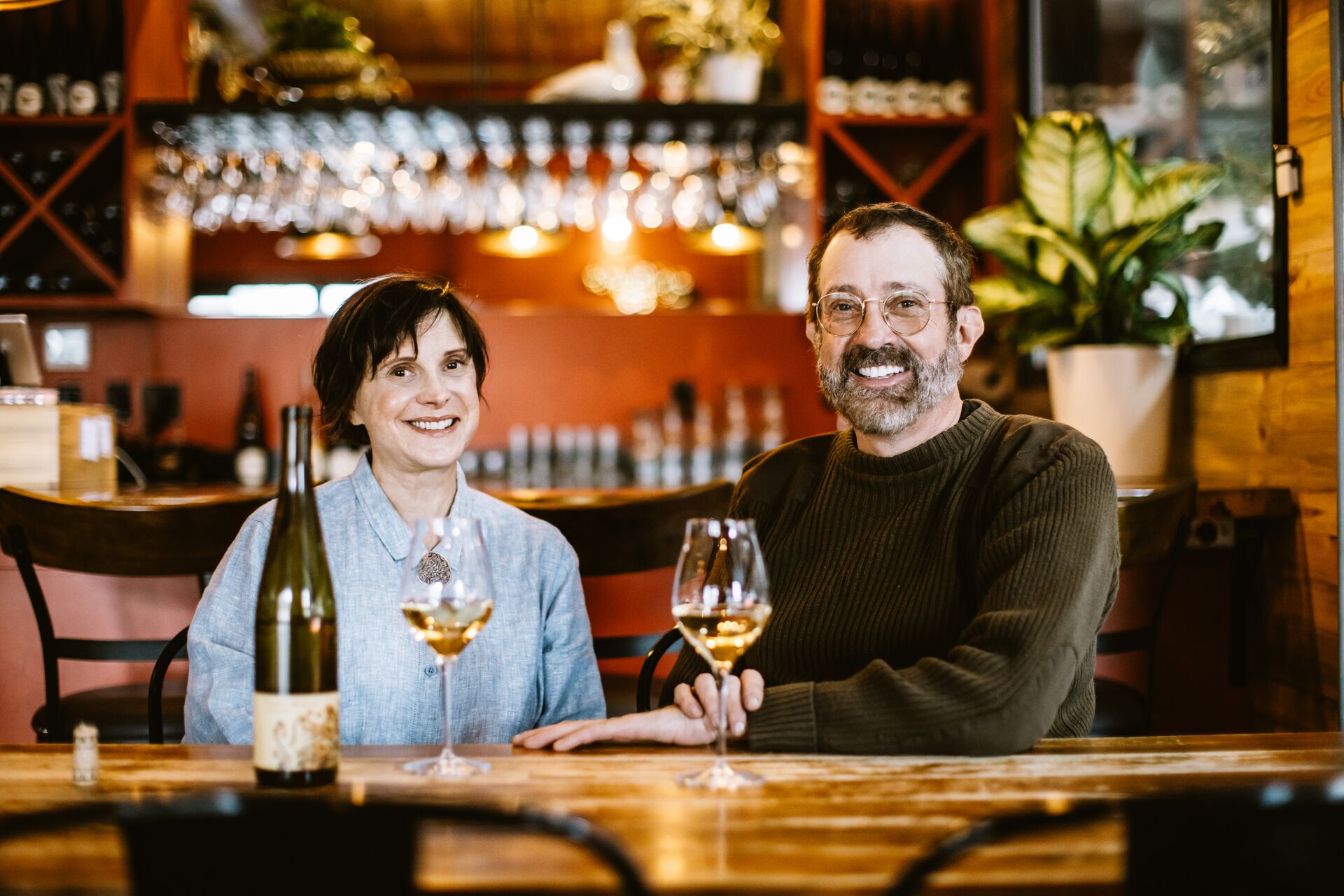 Olga and Barnaby smiling at the tasting room bar with glasses of white wine